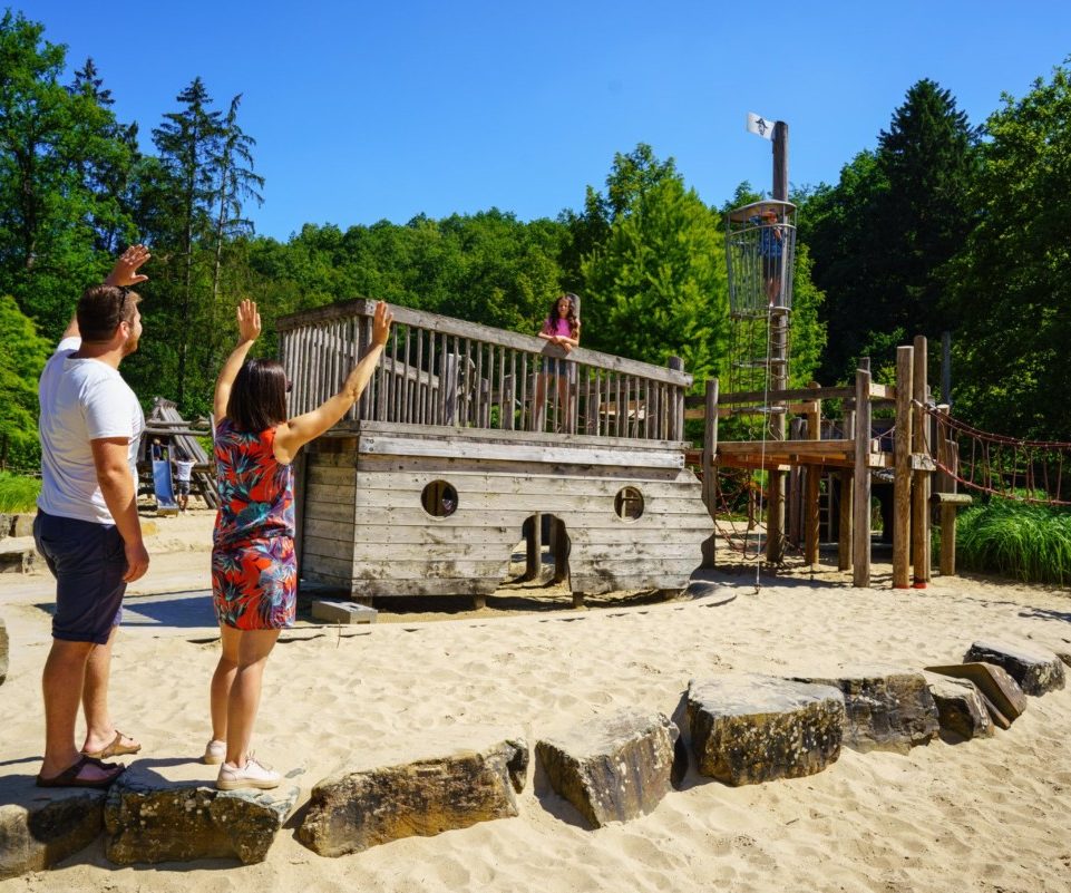 plaine-de-jeux-domaine-de-chevetogne Parents qui font signe à leur enfant qui joue sur une plaine de jeux du Domaine de Chevetogne lors d'une journée ensoleillée