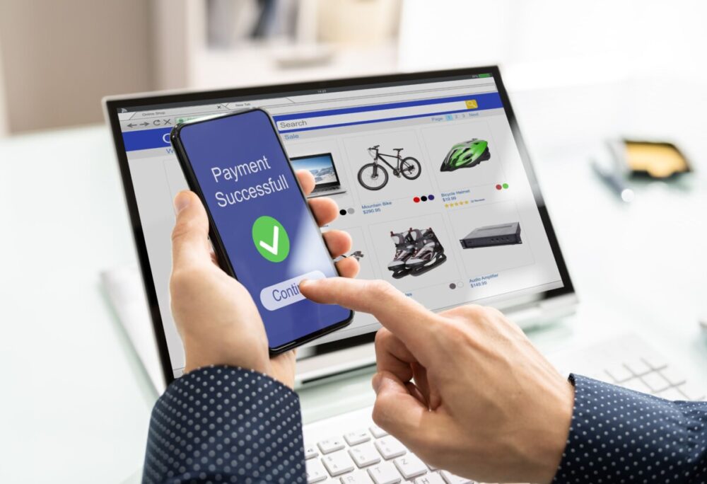 mains qui réalisent un paiement via un smartphone avec un ordinateur ouvert sur un e-shop en arrière-plan
