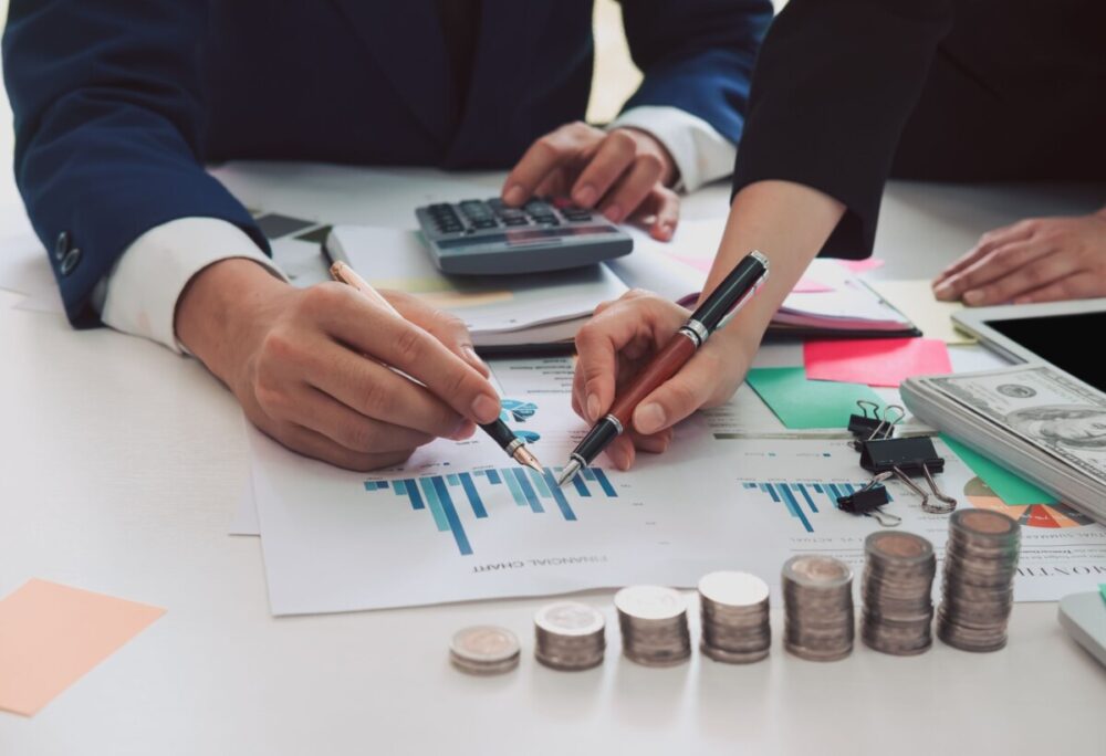 Deux personnes qui tiennent leur stylo en mains avec des piles de pièces devant eux