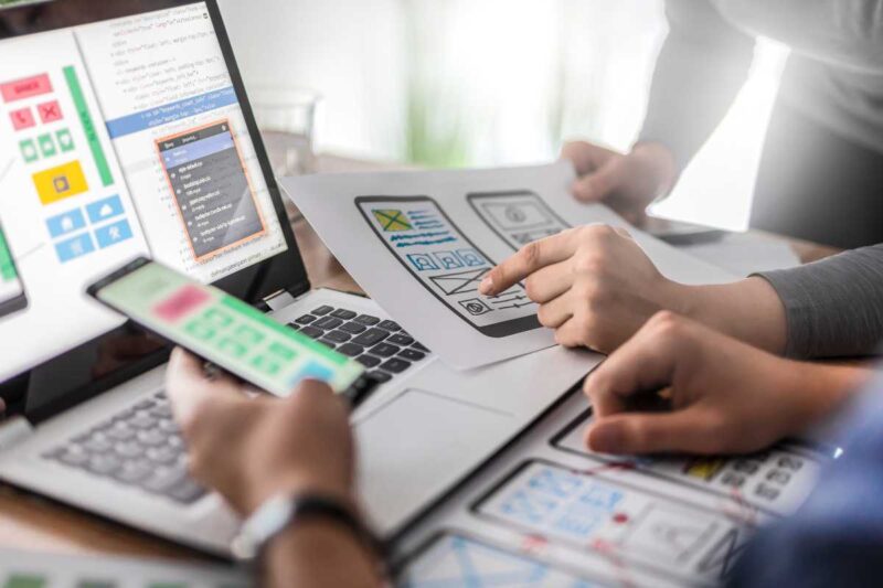 Deux personnes développent des interfaces utilisateur, testant des concepts sur un smartphone et un ordinateur dans un bureau éclairé.